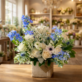 Arrangement of white roses and blue flowers in a burlap-wrapped square container on a wooden table