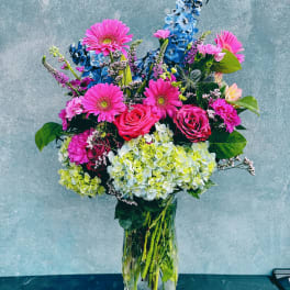 Bright mixed bouquet in a clear glass vase with pink gerberas, roses, hydrangeas, and blue delphinium.