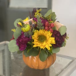 Fall floral arrangement in a pumpkin-shaped container with a sunflower and purple blooms