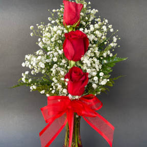 Three red roses in a clear vase with baby's breath and a red ribbon