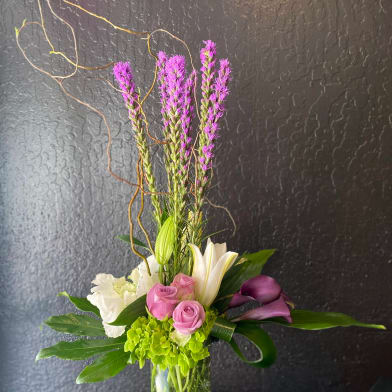 Tall vase arrangement with purple spikes, pink roses, white lilies, and dark purple calla lilies