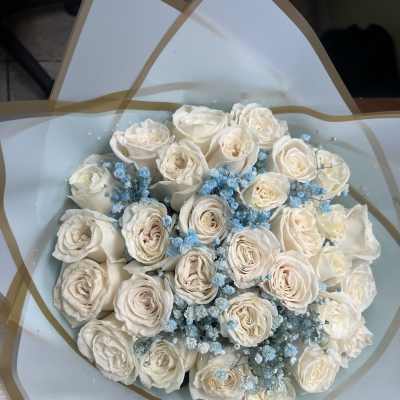 Round bouquet of cream roses with clusters of light blue filler flowers wrapped in pale blue paper.