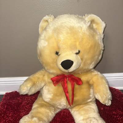 Large beige teddy bear with a red ribbon sitting on a red rug