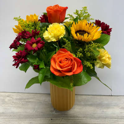 Mixed arrangement of orange roses, yellow carnations, red mums, and sunflowers in a tall gold vase