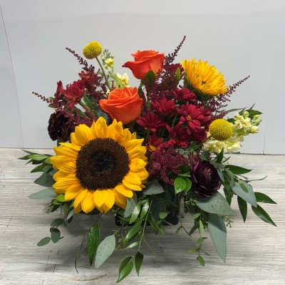 Low arrangement with a large sunflower, orange roses, red mums, and yellow accents in a black rectangular vase.