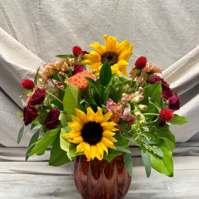 Mixed bouquet with sunflowers, red roses, and peach blooms arranged in a ribbed amber glass vase