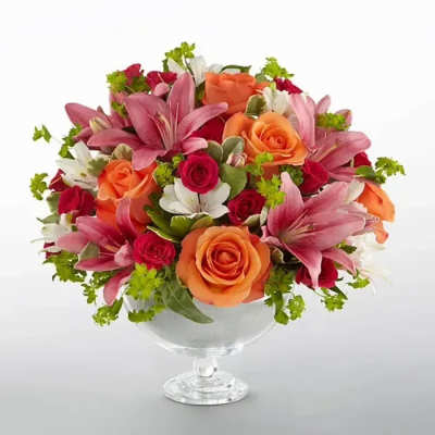 Low arrangement of pink lilies with orange and red roses in a clear glass pedestal bowl