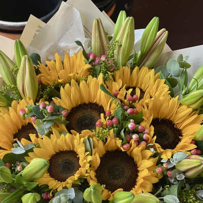 Hand-tied bouquet of yellow sunflowers with green lily buds and pink berries wrapped in cream paper.