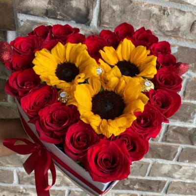 Heart-shaped box filled with red roses, three yellow sunflowers, and gold-wrapped chocolates.
