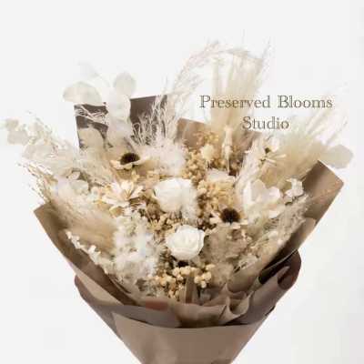 Neutral-toned bouquet of preserved white roses and dried flowers wrapped in brown paper with a dark ribbon.