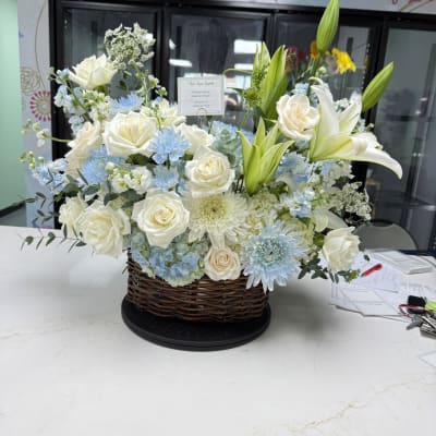 White roses and lilies arranged in a wicker basket with pale blue flowers.