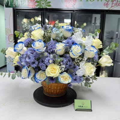 Basket arrangement of blue-edged white roses and purple flowers with a white ribbon