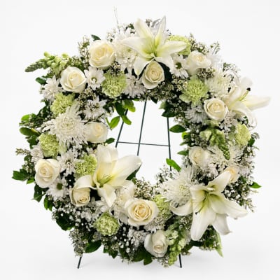 White floral wreath with roses, lilies, and green accents on a stand