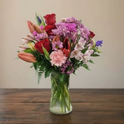 Mixed bouquet of roses, lilies, and purple flowers in a clear glass vase
