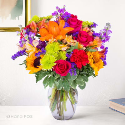 Bright mixed bouquet in a clear glass vase with roses, lilies, sunflowers, and purple accents