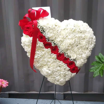 Standing heart-shaped white carnation wreath with diagonal red roses and a red ribbon bow on an easel