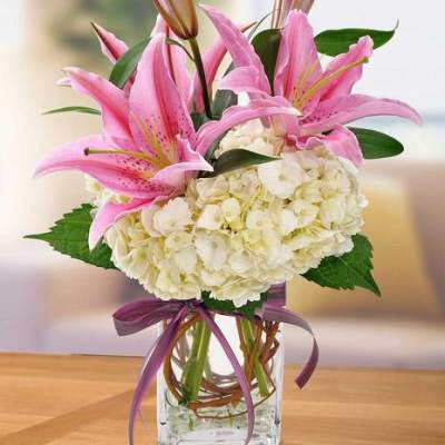 Pink lilies and white hydrangeas in a clear glass vase with a purple ribbon