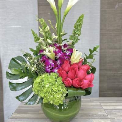 Tall arrangement of white calla lilies, pink roses, orchids, and green hydrangea in a round glass vase