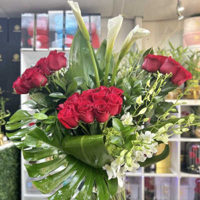Tall arrangement of red roses, white calla lilies, and orchids in a clear glass vase with large glossy leaves