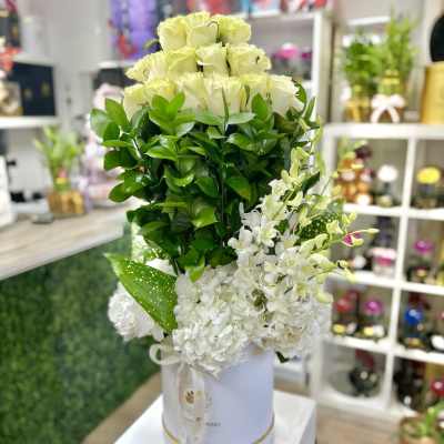 Tall arrangement of pale roses, white blooms, and dense greenery in a white hatbox container