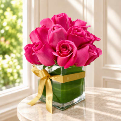 Pink roses arranged in a clear square vase with a gold ribbon