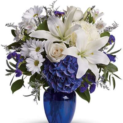 Mixed white lilies, roses, daisies, and blue hydrangeas arranged in a cobalt blue glass vase.