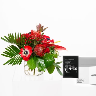 Low red and green floral arrangement in a clear glass vase with bold tropical leaves and a black greeting card beside it.