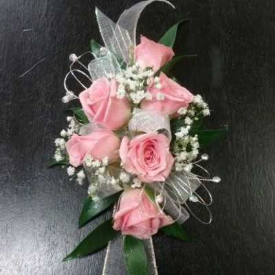 Pink rose wrist corsage with white filler flowers and sheer ribbon on a dark surface