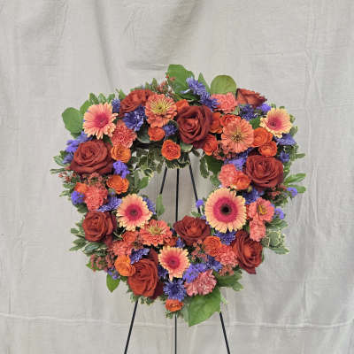 Heart-shaped standing wreath with red roses, orange gerbera daisies, and purple flowers on a black easel