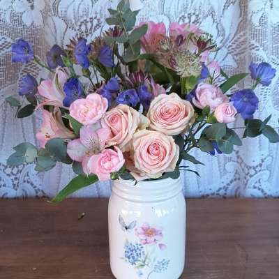 Pastel pink and blue flower arrangement in a white painted jar-style vase on a wooden surface