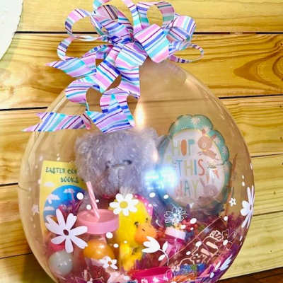 Clear balloon gift with a teddy bear, Easter treats, and a striped ribbon bow