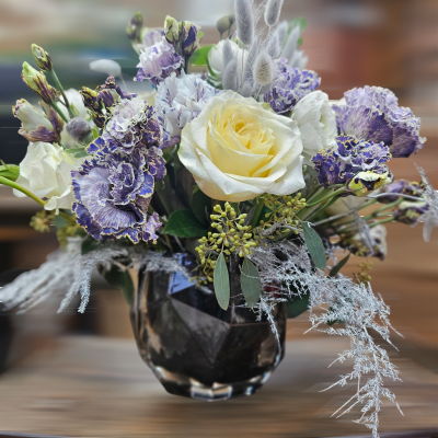 Short arrangement of cream roses and purple ruffled flowers in a dark glass vase
