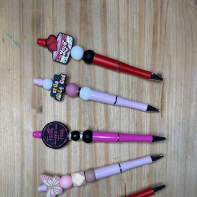 Five decorated pens with beads and charms arranged on a wooden surface.