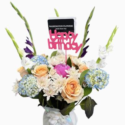 Birthday flower arrangement with peach roses, blue hydrangeas, and a pink Happy Birthday topper in a wrapped vase