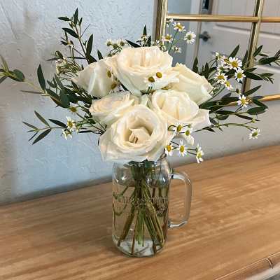 White rose and mini daisy arrangement in a clear mason jar on a wooden table.