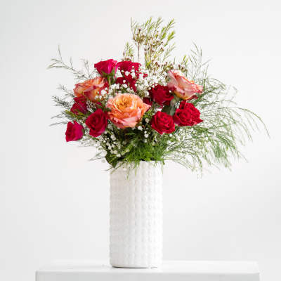 Tall arrangement of red and peach roses with white filler flowers in a textured white vase