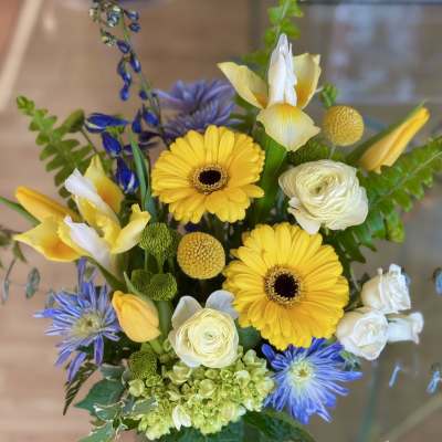 Bright mixed arrangement with yellow gerbera daisies, white blooms, and blue flowers in a green glass vase