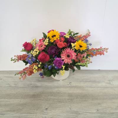 Low colorful arrangement of roses, gerbera daisies, sunflowers, and mixed blooms in a white ceramic bowl.
