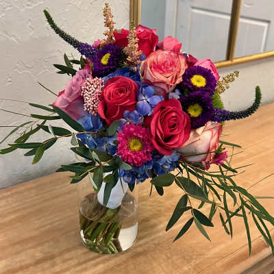 Bright mixed bouquet of pink roses, blue hydrangea, and purple mums in a clear glass vase