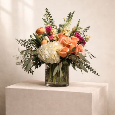Peach roses, white hydrangea, and colorful mixed flowers in a clear glass cylinder vase.
