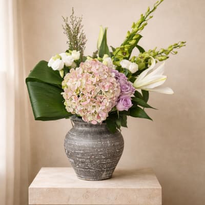 Asymmetrical arrangement of blush hydrangea, lavender roses, white lilies, and broad green leaves in a gray ceramic vase