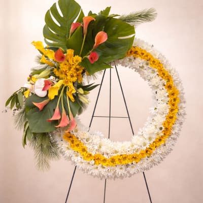 Large white and yellow standing wreath with pink-orange calla lilies and tropical foliage on a metal easel.
