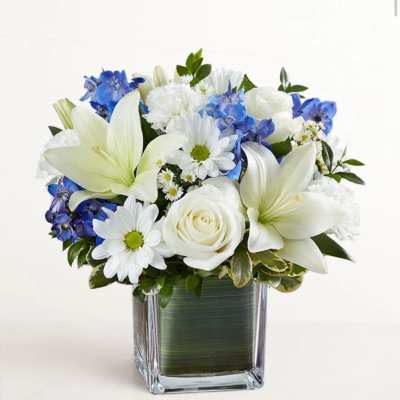White lilies, roses, and blue flowers arranged in a square glass vase