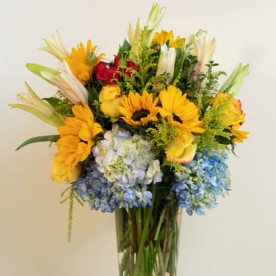 Mixed bouquet in a clear glass vase with sunflowers, roses, lilies, and hydrangeas