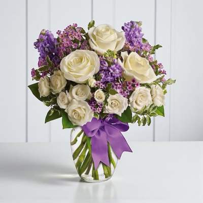 White roses and purple flowers in a clear glass vase with a purple ribbon