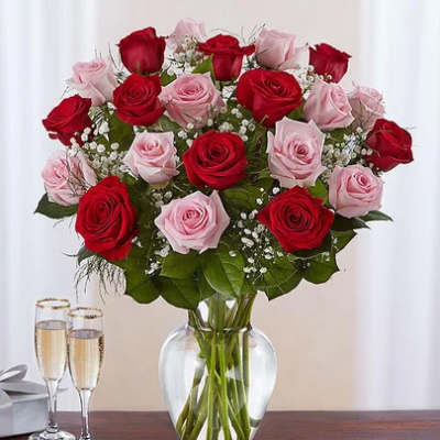 Arrangement of red and pink roses with baby's breath in a clear glass vase beside two champagne flutes.
