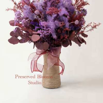 Tall bouquet of lavender and burgundy preserved flowers in a rope-wrapped vase with a pink ribbon bow