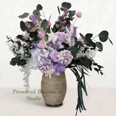 Tall arrangement of lavender and white preserved flowers with dark greenery in a textured beige vase