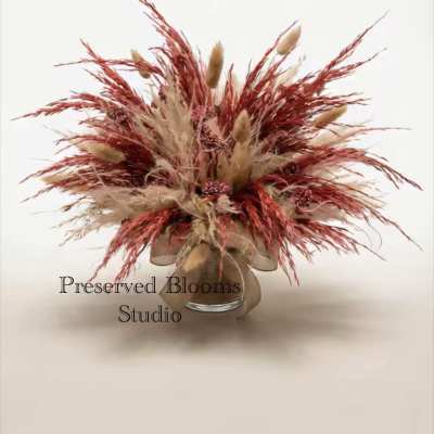 Preserved arrangement of tan and rust dried grasses and seed clusters in a clear glass vase.