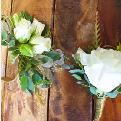 Two white rose boutonnieres with greenery on a wooden surface